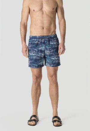 Hombre sin camisa que lleva puestos pantalones de baño azul marino con estampado y sandalias negras con hebilla, de pie sobre un fondo gris claro.