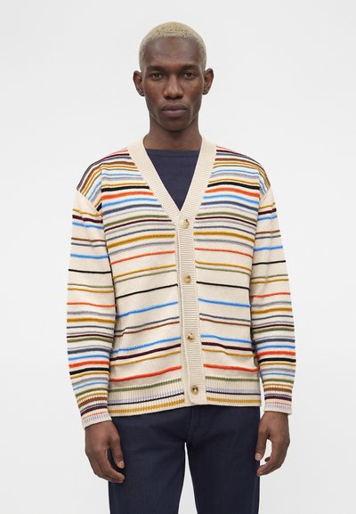 Μαύρος άνδρας με ξανθά κοντά μαλλιά, που φοράει πολύχρωμη ριγέ ζακέτα με κουμπιά και σκούρο παντελόνι πάνω σε απλό λευκό φόντο.