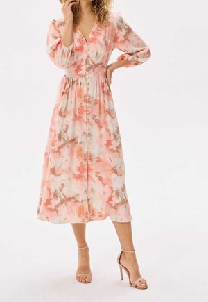 Kvinde iført en lyserød og cremefarvet blomsterprint midi-kjole med lange pufærmer og knaplukning foran, kombineret med nøgenfarvede høje ankelrem hæle.