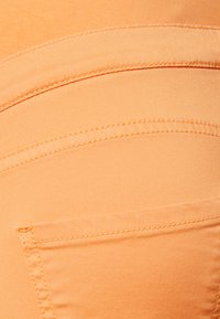 Têxtil laranja com textura suave, apresentando costuras curvas e um único bolso traseiro com detalhes de costura visíveis.