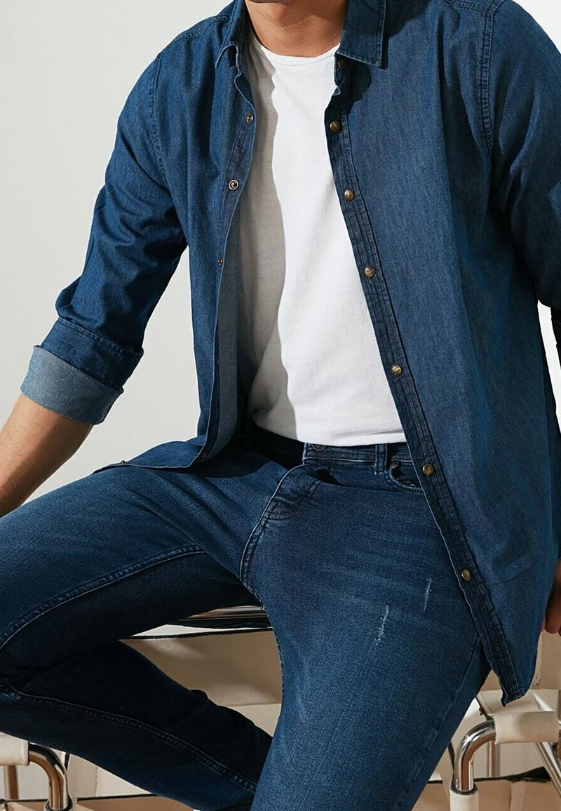 Homme portant une chemise en jean bleue et un jean avec un t-shirt blanc, assis avec un bras reposant sur une chaise.