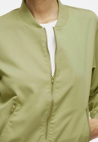 Giacca bomber verde chiaro con colletto a costine, zip frontale e tasche laterali. Realizzata in tessuto liscio con un design semplice e pulito.