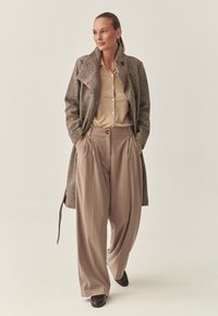 Pruun tweed ülikond üle beeži nööpidega särk, koos beežide laia säärega pükstega. Mustad kingad täiendavad riietust. Minimalistlik disain ja tekstuurid.