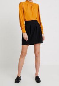 Femme portant un chemisier à manches longues moutarde avec un col smocké, une jupe plissée noire et des mocassins noirs, debout devant un fond uni.