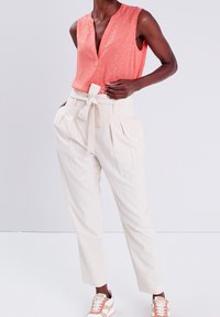 Pfirsichfarbendes ärmelloses Oberteil mit silbernem Muster, kombiniert mit cremefarbenen plissierten Hosen mit Bindegürtel. Weiße und beige Sneakers runden den Look ab.