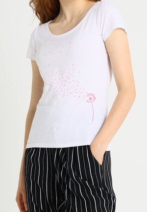 Kobieta ubrana w biały t-shirt z różowym wzorem dmuchawca i czarne spodnie z białymi pionowymi paskami, ręka w kieszeni.