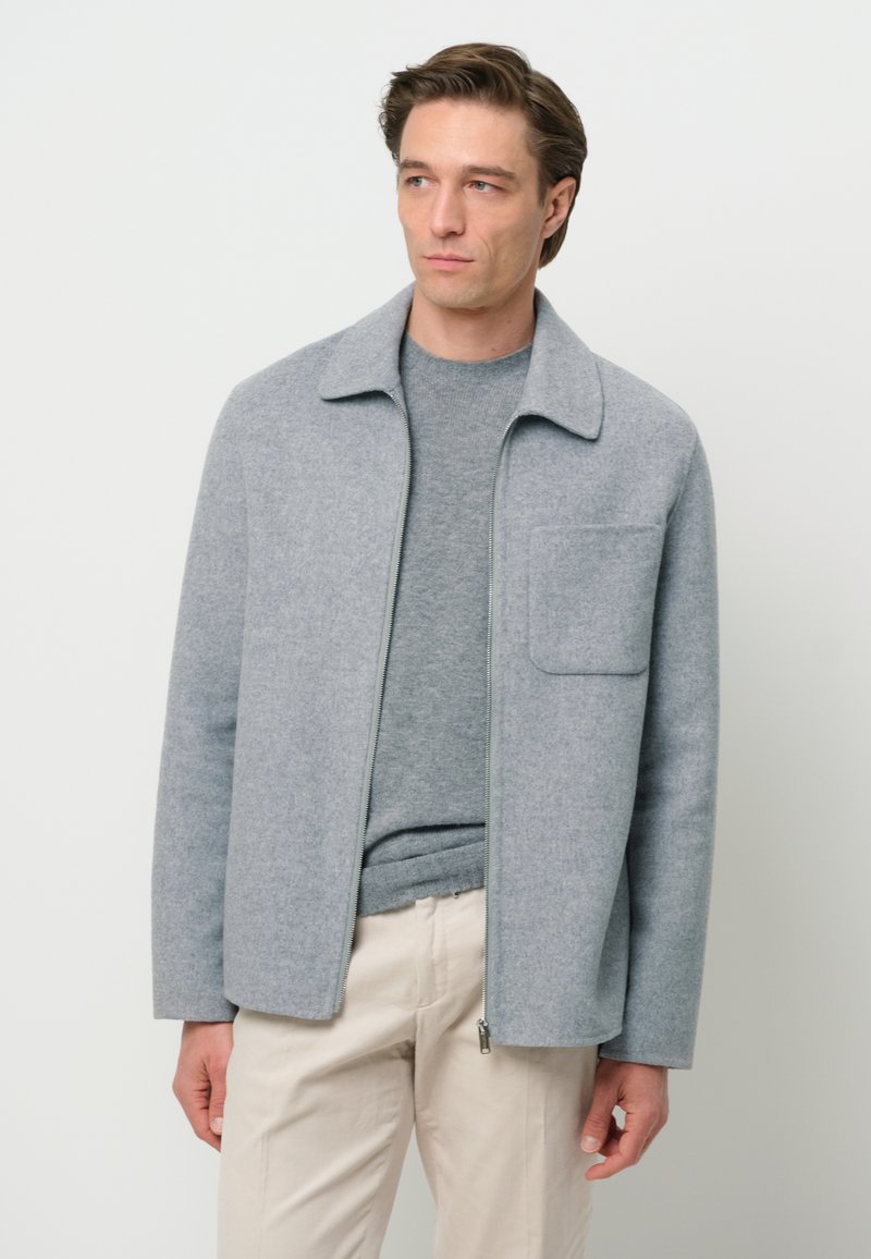 Giacca grigia in blend di lana con zip frontale, colletto a punta e taschino sul petto, indossata sopra un maglione grigio e pantaloni beige. Texture liscia.