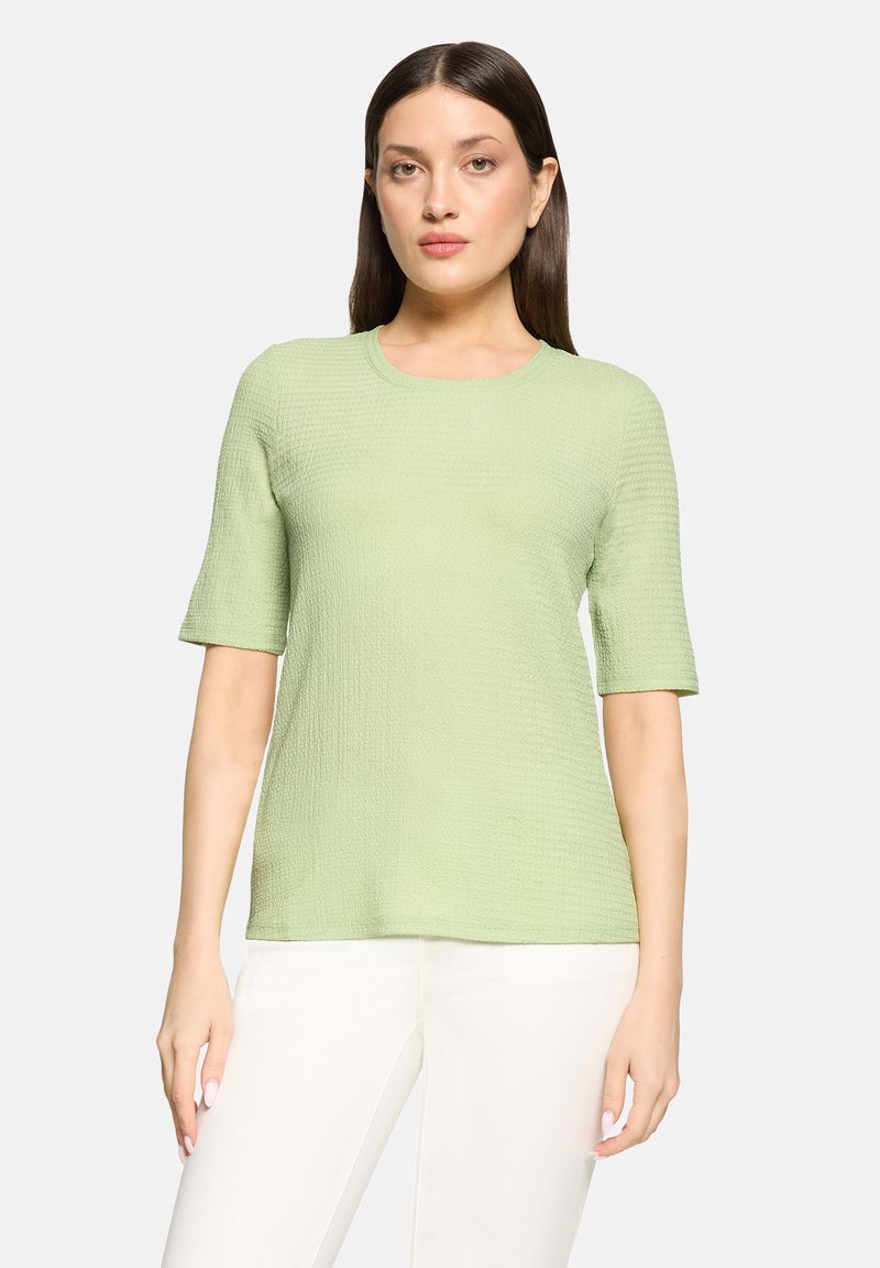 Kobieta o długich ciemnych włosach, ubrana w jasnozielony, teksturowany top z krótkim rękawem i białe spodnie, stojąca na jednolitym tle.