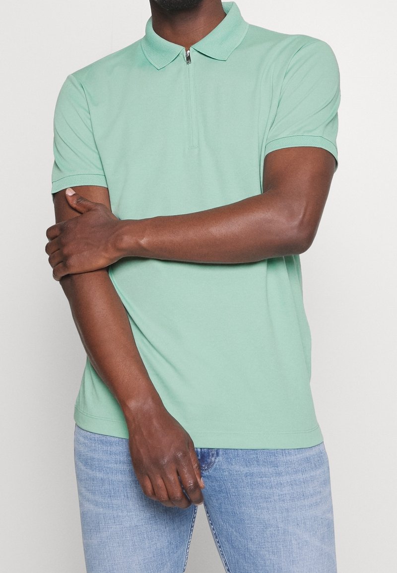 Polo shirt vert clair à manches courtes, avec un col zippé et une texture lisse, associé à un jean bleu clair. Design minimaliste.