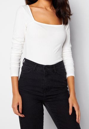 Mujer con una camiseta blanca de manga larga con cuello cuadrado metida en unos vaqueros negros de talle alto, sobre un fondo liso.