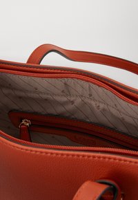 Sac à main en cuir orange brûlé avec une texture lisse, doté d'une poche à fermeture éclair et d'une doublure intérieure à motif. Poignées courtes incluses.