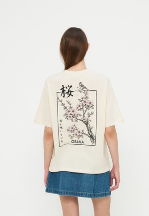Young woman wearing a cream t-shirt with a pink cherry blossom and bird illustration and the word Osaka on the back.