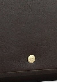 Bolso de cuero marrón con una textura suave y un botón circular dorado con un logo. El diseño es minimalista y funcional.