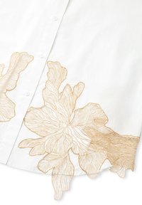 Camisa blanca con un diseño floral bordado en oro a lo largo del dobladillo, con detalles de botones y una textura suave y ligera.