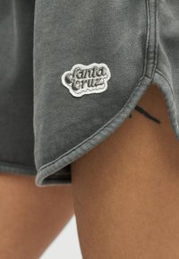 Γκρι σορτς με καμπυλωτό τελείωμα και λευκή ετικέτα λογότυπου «Santa Cruz» που φοριέται στο μηρό ενός ατόμου, με ένα μικρό μαύρο τατουάζ να διακρίνεται μερικώς.