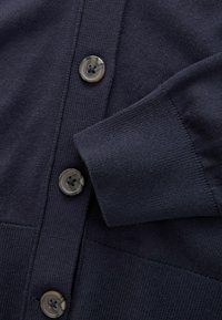 Närbild av en mörk marinblå stickad cardigan med tre rundade marmorerade knappar och en ribbad mudd på ärmen.