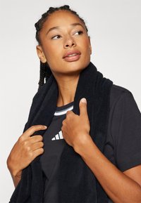 Μαύρη πετσέτα κρεμασμένη γύρω από τον λαιμό ενός ατόμου που φοράει σκούρο πουκάμισο με λευκό λογότυπο. Το πουκάμισο διαθέτει αντίθεση στη γραμμή του λαιμού με ρίγες.