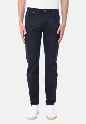 Uomo che indossa jeans slim fit blu scuro e sneaker bianche, in piedi con le braccia rilassate lungo i lati contro uno sfondo bianco.