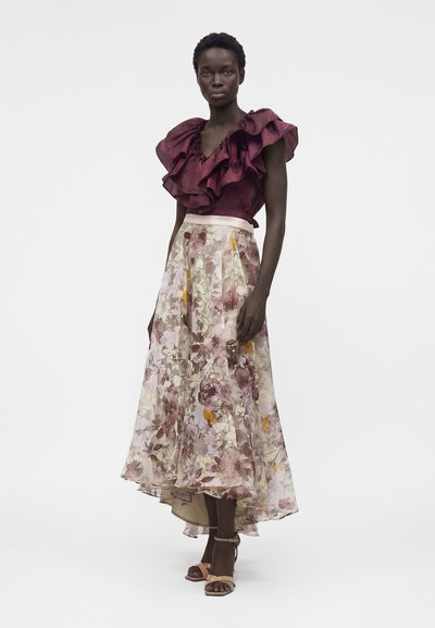 Γυναίκα που φοράει μπορντό αμάνικη μπλούζα με βολάν και φούστα με λουλουδένιο μοτίβο σε ροζ και κίτρινους τόνους, συνδυασμένη με γόβες με λουράκι στον αστράγαλο σε γυμνό χρώμα.