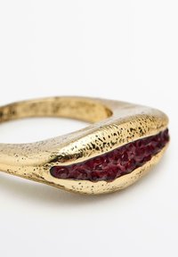 Anello dorato con superficie texturizzata, caratterizzato da un'incrustazione cristallina rossa disposta in una forma curva e organica.