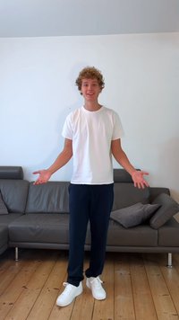 En person i en vit t-shirt och mörka byxor står med armarna utsträckta på ett trägolv nära en grå lädersoffa med kuddar.