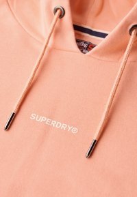 Hoodie w kolorze brzoskwiniowym z regulowanym sznurkiem zakończonym metalowymi końcówkami oraz białym logo „SUPERDRY”. Miękka tekstura, casualowy styl, standardowe dopasowanie.