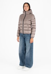 Licht taupe pufferjack met een hoge kraag, een rits aan de voorkant en elastische manchetten, gecombineerd met losse blauwe spijkerbroeken en witte sneakers.