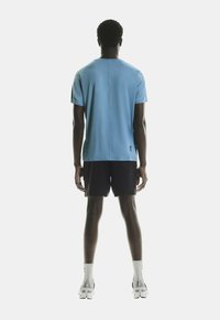 T-shirt sportif bleu clair à manches courtes, associé à un short noir, vu de l'arrière. Présente des coutures minimales et un logo près de l'ourlet.