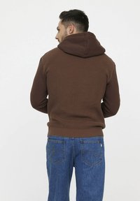 Brauner Kapuzenpullover mit strukturiertem Muster und geripptem Saum, kombiniert mit blauen Jeans mit Gesäßtaschen und Steppnähten.