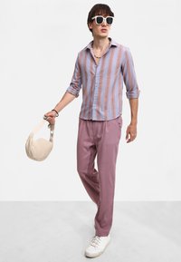 Camisa ligera a rayas en azul y rosa, combinada con pantalones de color púrpura claro. Lleva un bolso beige, unas gafas de sol blancas y zapatillas.