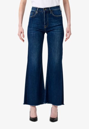 Donna che indossa jeans a gamba larga blu scuro con orli grezzi e tacchi alti neri a punta, abbinati a una maglietta bianca.