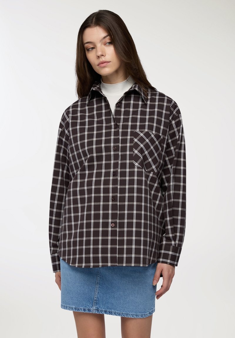 Camicia a quadri marrone e bianca con maniche lunghe, colletto, chiusura con bottoni e una tasca sul petto. Abbinata a una gonna di jeans azzurri chiari.