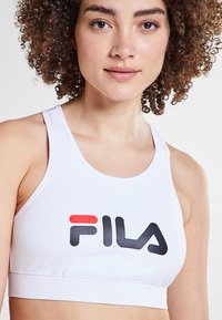Sujetador deportivo blanco con el logo de Fila en rojo y negro. Presenta un diseño simple y corto, además de una tela suave y elástica. Con tirantes ajustables.
