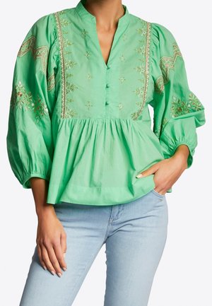 Zielona bluzka typu peplum ze złotym haftem, zapinanym dekoltem w kształcie litery V i balonowymi rękawami, noszona z jasnoniebieskimi dżinsami.