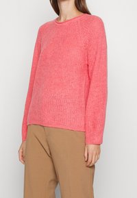 Helles Korallpullover mit strukturiertem Strick, Rundhalsausschnitt und langen Ärmeln. Über beigen weiten Hosen getragen.