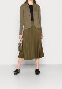 Olive green fitted blazer with two pockets, paired with a pleated olive green midi skirt. Black shoes and a small handbag complete the look.