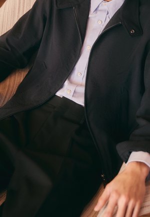Dunkelgraue Zip-Jacke aus strukturiertem Stoff, hellblaues kariertes Hemd und schwarze plissierte Hose, die glatte Oberflächen und maßgeschneiderte Gestaltung zeigen.