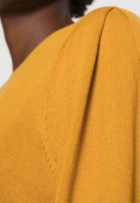 Närbild av en person som bär en senapsgul stickad tröja med detaljerad sömnad på axelpartiet.