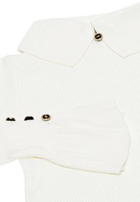 Top bianco a costine con colletto classico e maniche a palloncino. I dettagli con bottoni dorati ai polsini delle maniche aggiungono un tocco di eleganza. Tessuto dalla texture morbida.