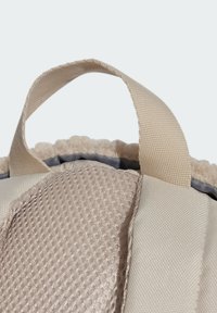 Detalle de una mochila de tela beige que muestra un panel de malla texturizada, bordes lisos y un asa plana en la parte superior para opciones de transporte.
