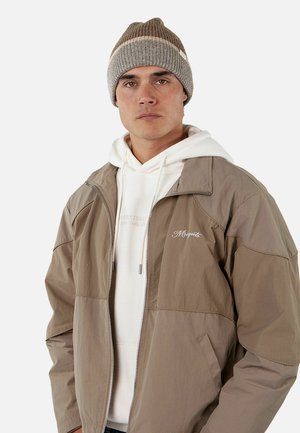 Chaqueta ligera color marrón con cierre de cremallera, sudadera blanca debajo y gorro de punto marrón. Logo simple en la chaqueta. Textura suave.