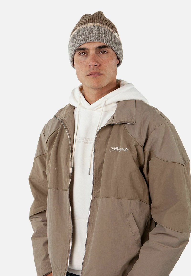 Giacca leggera tan con chiusura a zip, felpa bianca sotto, e berretto lavorato a maglia marrone. Logo semplice sulla giacca. Texture liscia.