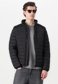 Black puffer jacket with horizontal quilted design, high collar, and zippered front. Worn over a grey shirt, complemented by sunglasses.