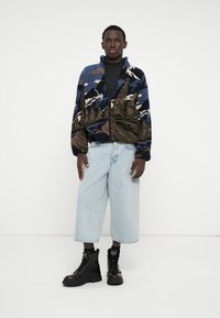 Puszysta kurtka zapinana na zamek w niebiesko-zielono-beżowym wzorze górskim; zestawiona z jasnoniebieskimi, szerokimi dżinsami i czarnymi sznurowanymi butami.