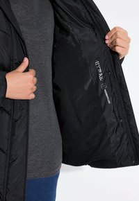 Chaqueta acolchada negra con un exterior brillante, que cuenta con un bolsillo interior con cremallera y etiqueta de marca. El interior es suave y liso.