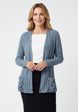 Kobieta o blond włosach, nosząca kolczyki koła, jasnoniebieski prążkowany kardigan z przednimi kieszeniami, biały top i czarną spódnicę, uśmiechająca się do aparatu.