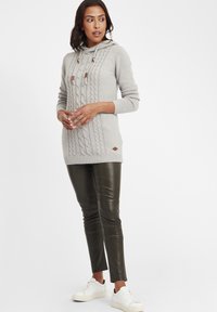 Hellgrauer Strickpullover mit Kapuze, ausgestattet mit braunen Kordel-Akzenten. Kombiniert mit dunkelgrünen, enganliegenden Hosen und weißen Turnschuhen.