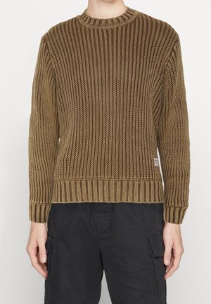 Person trägt einen braunen, gerippten Strickpullover mit langen Ärmeln kombiniert mit schwarzen Cargo-Hosen vor einem schlichten Hintergrund.