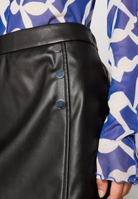 Mustat nahkahameet, joissa on sileä pinta ja hopeiset nepparit sivussa. Sinivalkoraidallinen läpikuultava paita on osittain näkyvissä.