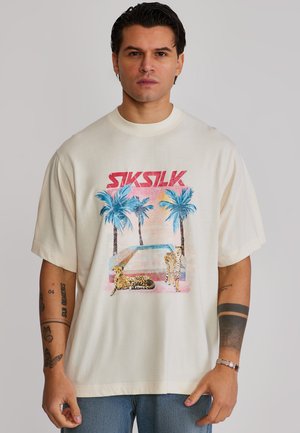 Muž nosiaci biele prehnane veľké tričko s farebnou grafikou palmových stromov, dvoch gepardov a červeným nápisom "SikSilk" na prednej strane.